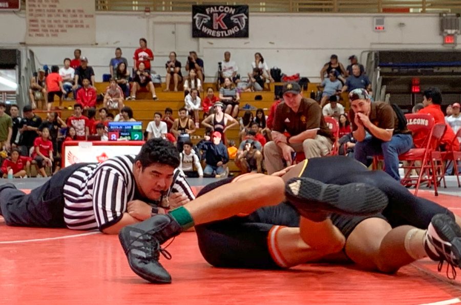 Ikaika Nishitomi referees a wrestling match on Mat 3 between wrestlers from Mililani (wearing gold on his left shoulder) and Campbell (wearing orange on his thigh) on Dec. 7 at Kalani High School. In the background, two Mililani coaches yell shots to their school’s wrestler, watching him drive his opponent for a pin. The Kalani Falcons hosted this season’s first wrestling tournament. Thirty-one Falcons participated: 17 junior varsity players and 14 varsity players. For this Round Robin tournament, wrestlers competed in three rounds per match. High schools participating in the competitions take turns hosting the winter sport’s nearly weekly Saturday events. If you’re interested in supporting Kalani’s wrestling team, visit the school’s athletics website for the tournament schedule. Photo & caption by Saara Nicole Chadwick. 